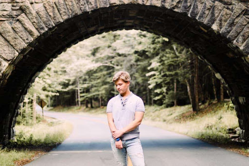 A photo of a well dressed blond young man standing underneath the arch of a bridge. The photo is very tasteful, and behind the boy is a vibrant green forest. He's standing on a path that winds through the woods.